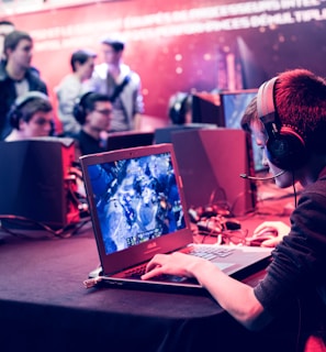 Several people are seated and focused on computer screens, wearing headsets, in an environment that appears to be a gaming tournament or esports event. The room is dimly lit with neon lighting, highlighting screens and creating an intense atmosphere.